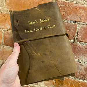 Hand holding up a brown leather rustic wrap journal with gold embossed title