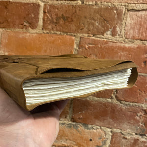 Hand holding a leather rustic wrap journal on its side against a brick background.