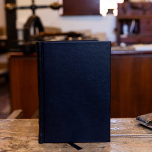 Small SB Libris Classic Journal in black leather, upright on a wooden bench with a subtle grain and traditional raised bands on the spine.