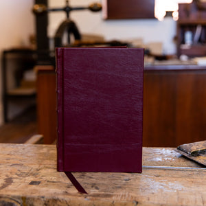Compact SB Libris full leather journal in maroon, photographed upright on a workbench with natural lighting and a matching ribbon.