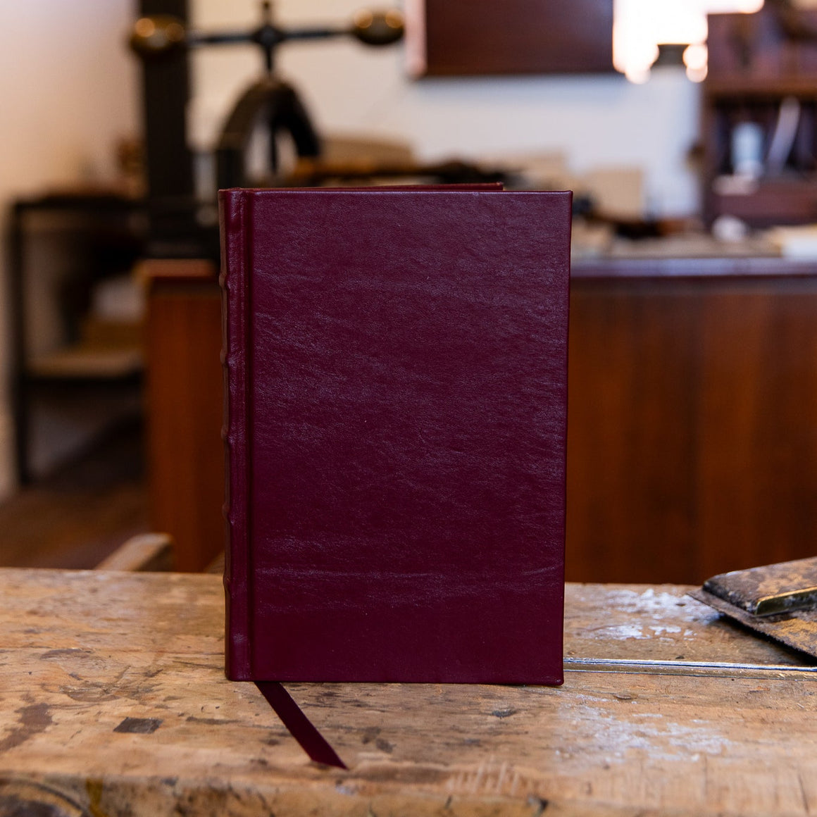 Compact SB Libris full leather journal in maroon, photographed upright on a workbench with natural lighting and a matching ribbon.