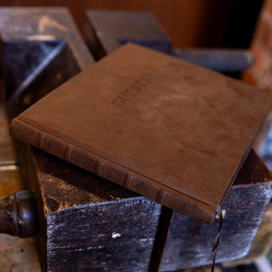 Aged goatskin leather guest book resting flat on a bookbinder’s clamp, showing raised spine bands and brown ribbon marker.
